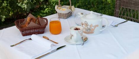 A breakfast table set outdoors with a basket of bread, a glass of orange juice, a teapot, and a cup of coffee.