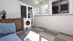 A laundry room with a washing machine, a drying rack, a potted plant, and a window with a view of buildings. - Apartment Ilinovic (Rovinj)
