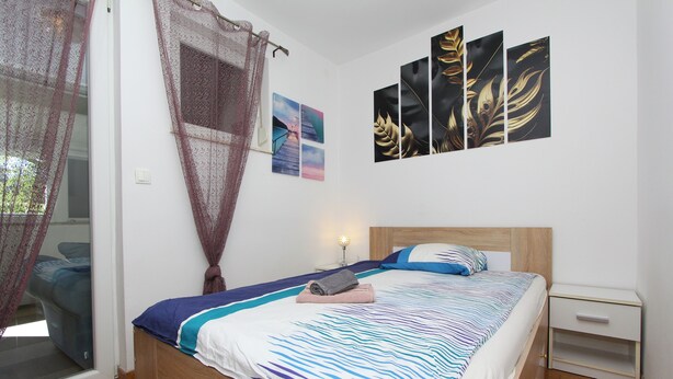 A bedroom with a bed, a nightstand, a wall with artwork, and a window with curtains. - Apartment Ilinovic (Rovinj)