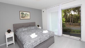 Quarto com uma cama grande, uma mesa de cabeceira, um abajur, um quadro na parede e vista para árvores através de uma porta de vidro deslizante.
