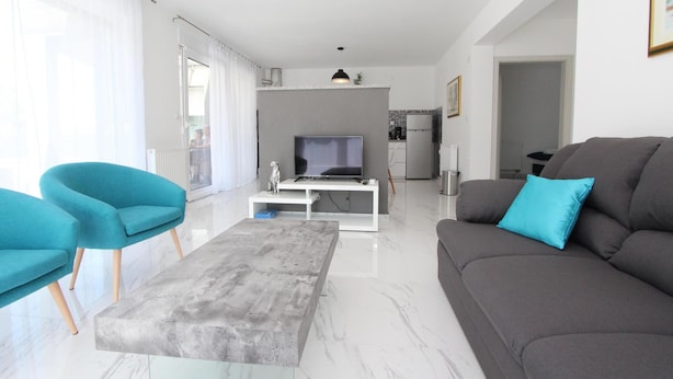 Uma sala de estar moderna com um sofá cinza, poltronas verde-água e uma mesa de centro cinza. Uma televisão de tela plana está montada em um aparador branco.