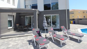 Uma área moderna de piscina ao ar livre com espreguiçadeiras e uma mesa, cercada por um edifício com grandes janelas e um terraço coberto.