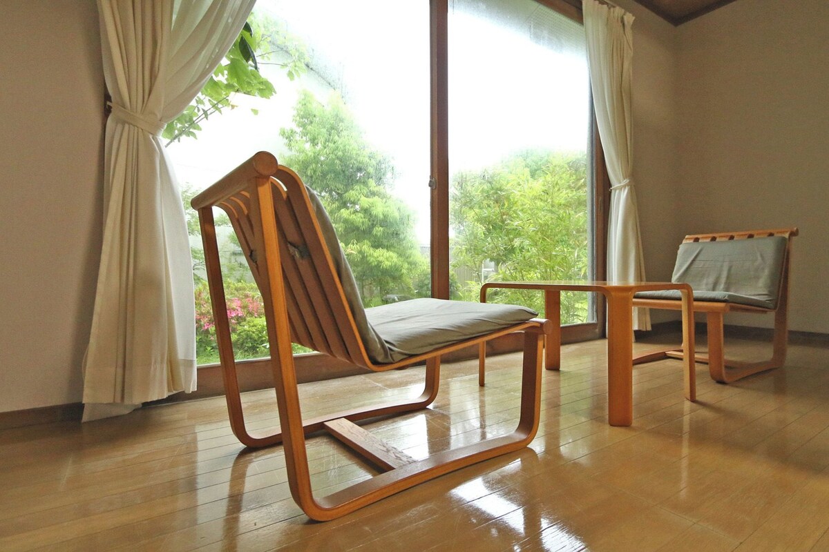 Una habitación con piso de madera, dos sillas de madera, un ventanal con cortinas y vistas a zonas verdes.