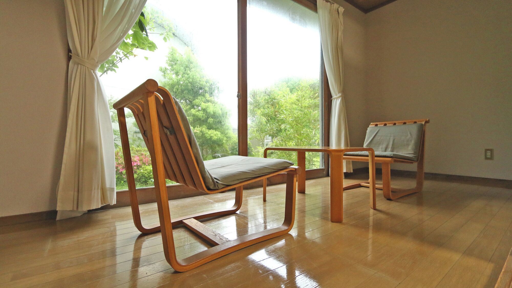 Una habitación con piso de madera, dos sillas de madera, un ventanal con cortinas y vistas a zonas verdes.