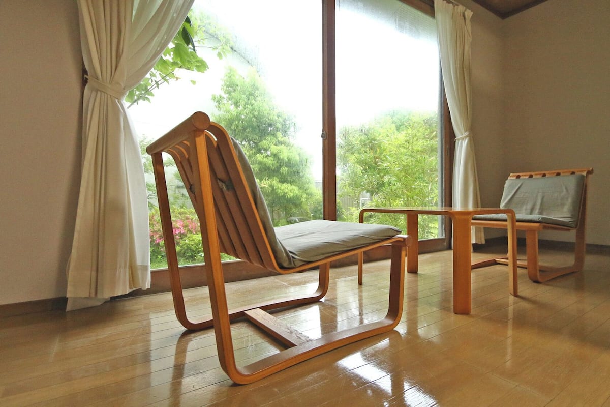 A room with wooden flooring, two wooden chairs, a window with curtains, and a view of greenery outside.