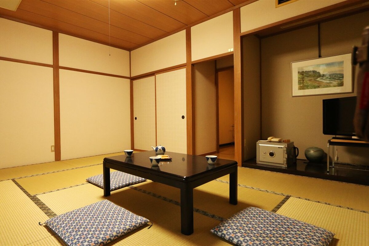 Une pièce avec un sol recouvert de tatamis, une petite table basse, des coussins, un téléviseur et un coffre-fort.