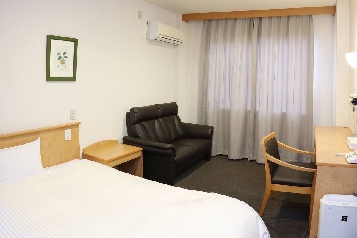 Une chambre d’hôtel comprenant un lit, un fauteuil en cuir, un bureau avec une chaise, un appareil de climatisation, un tableau encadré au mur et une fenêtre avec des rideaux.