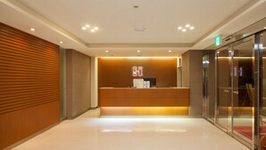 A modern reception area with a wooden reception desk, a glass elevator, and a clean, tiled floor. - Business Hotel Isesaki Heisei INN (Isesaki)