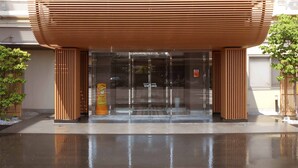 A modern building entrance with a wooden slatted canopy, glass doors, and a wet ground reflecting the structure. - Business Hotel Isesaki Heisei INN (Isesaki)