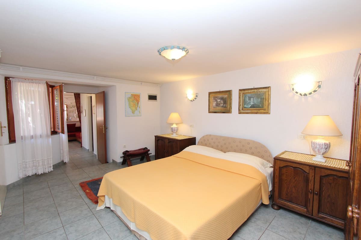 A bedroom with a large bed, two bedside tables with lamps, a wooden wardrobe, and a door leading to another room.