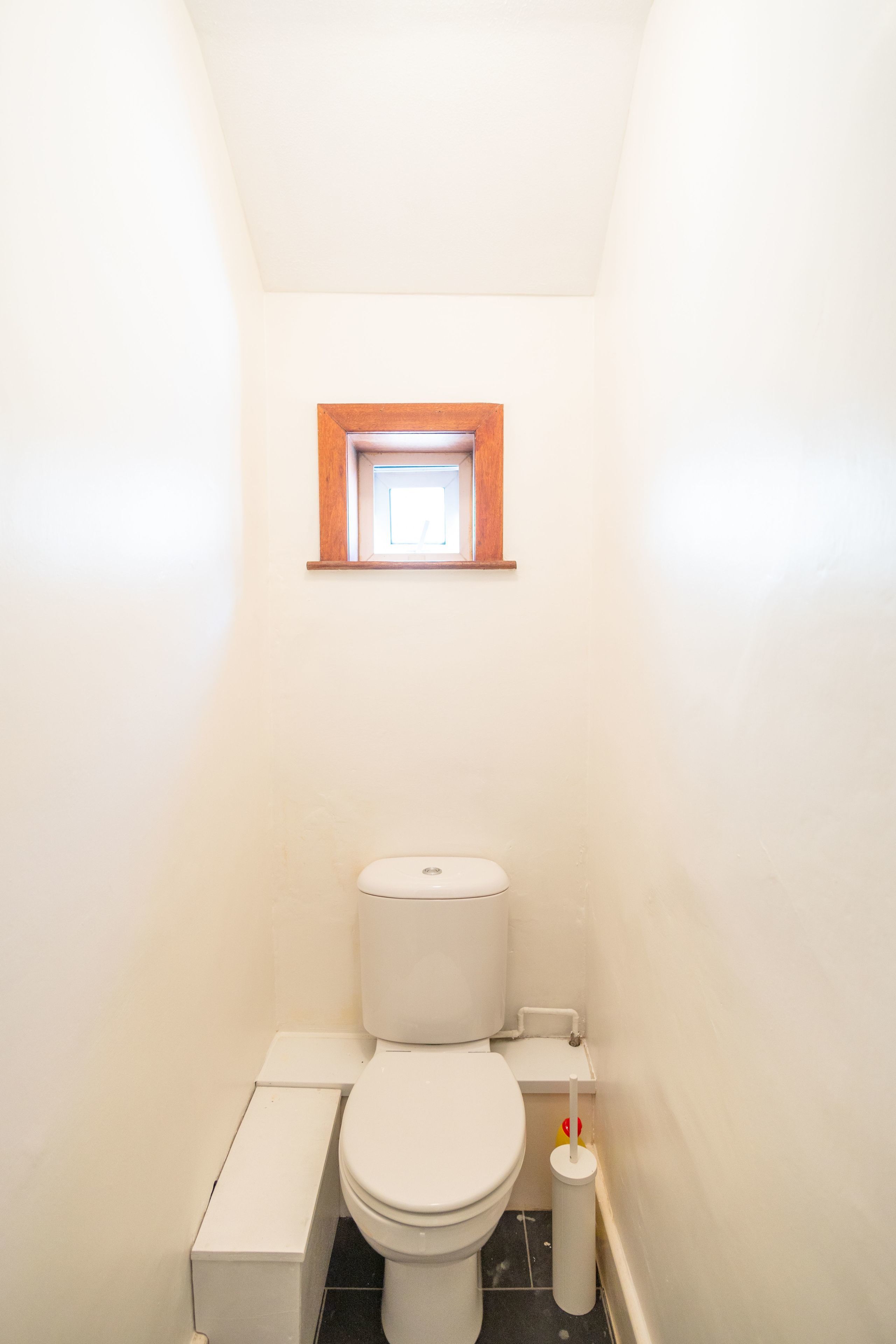 A compact bathroom with a toilet, a small window, and a toilet brush.