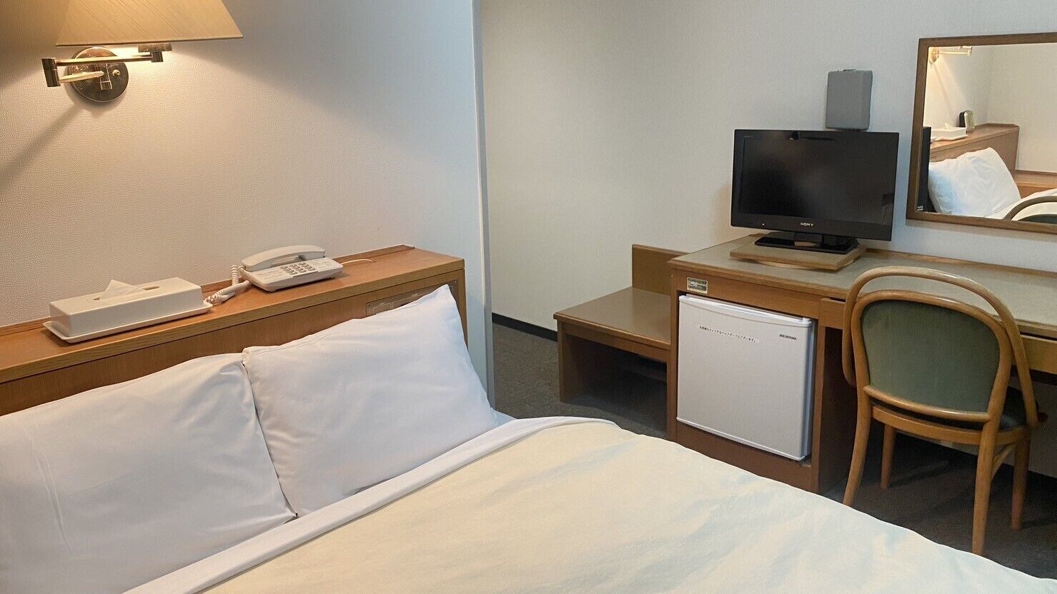 A hotel room with a bed, a desk with a television, a phone, a mini-fridge, and a chair.