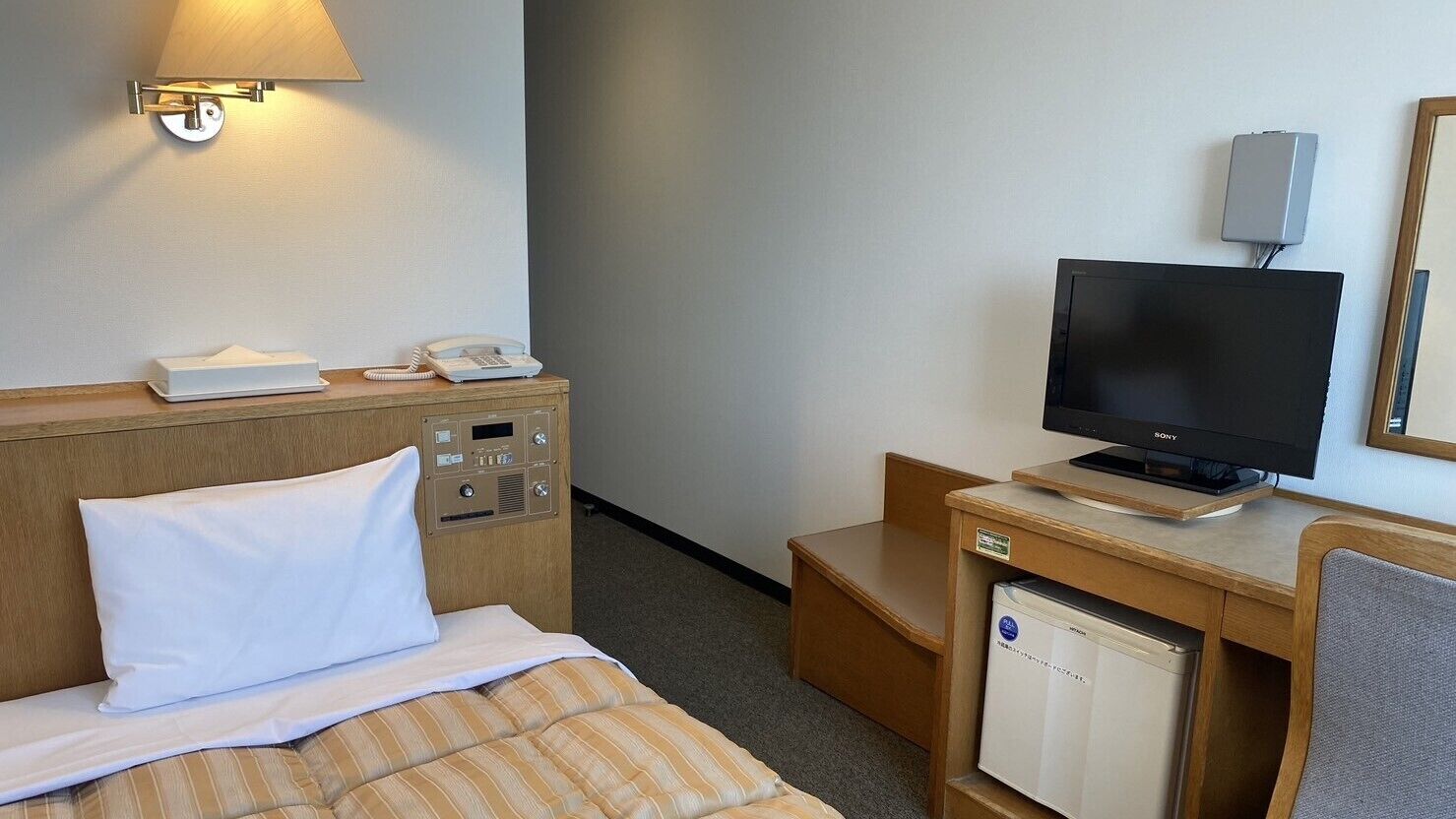 A hotel room with a bed, a desk with a television, a telephone, and a small refrigerator.