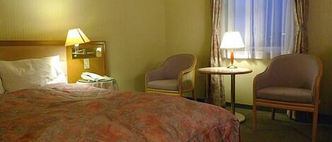 Une chambre d’hôtel avec un lit, deux chaises, une petite table avec une lampe, une table de chevet avec un téléphone et une fenêtre avec des rideaux.