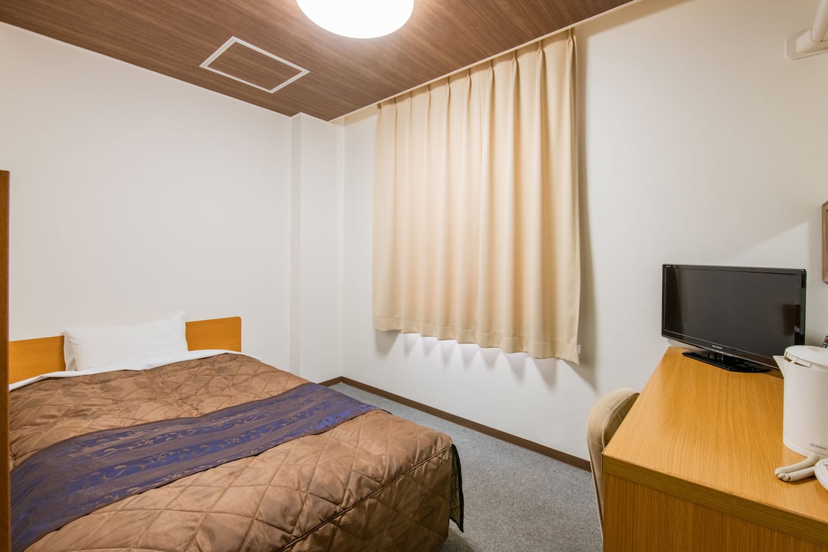 A hotel room with a bed, a desk with a television, and a window with curtains.