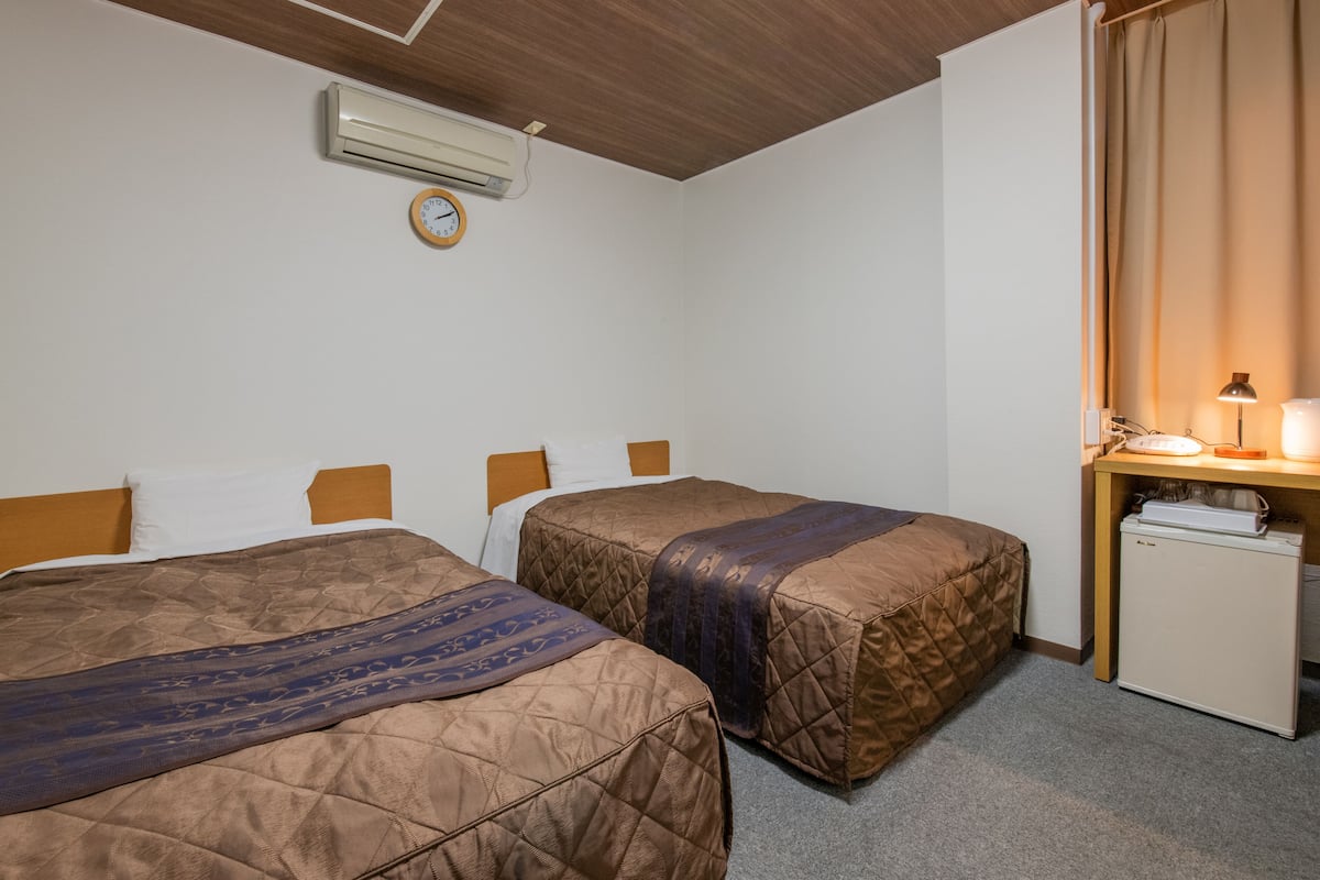A hotel room with two beds, a small desk with a lamp, an air conditioner, and a clock on the wall.