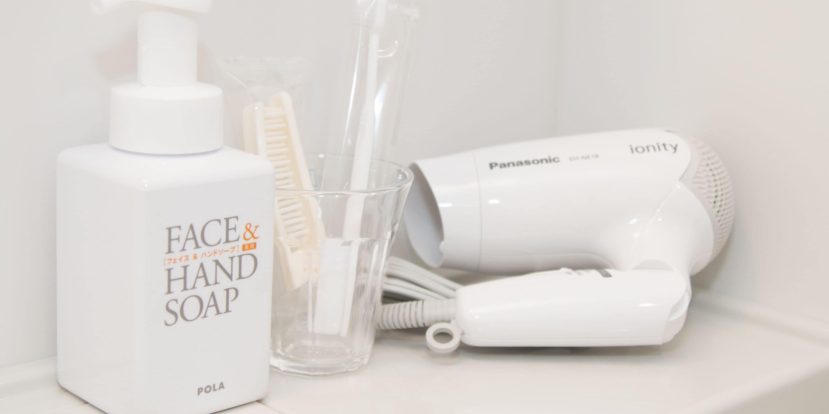 A bathroom with a bottle of face and hand soap, a hairdryer, and a toothbrush holder.
