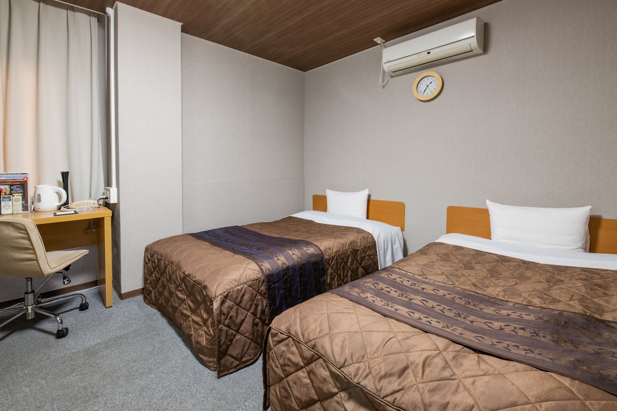 A hotel room with two beds, a desk, a chair, an air conditioner, and a clock on the wall.