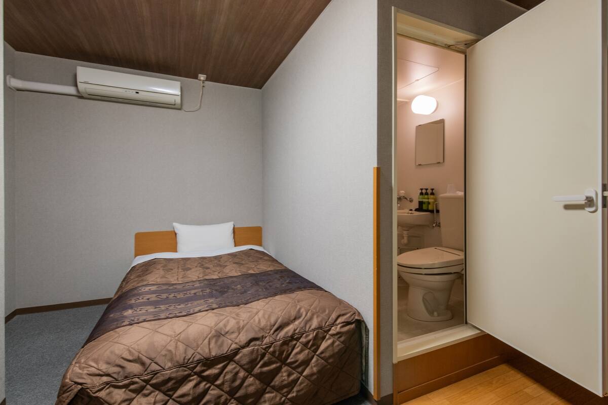 A bedroom with a bed, a wooden ceiling, a white wall, and a bathroom visible through an open door.
