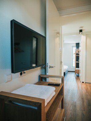 A modern hotel room with a wooden bed, a flat-screen TV mounted on the wall, and a hallway leading to a bathroom with a wooden bench.