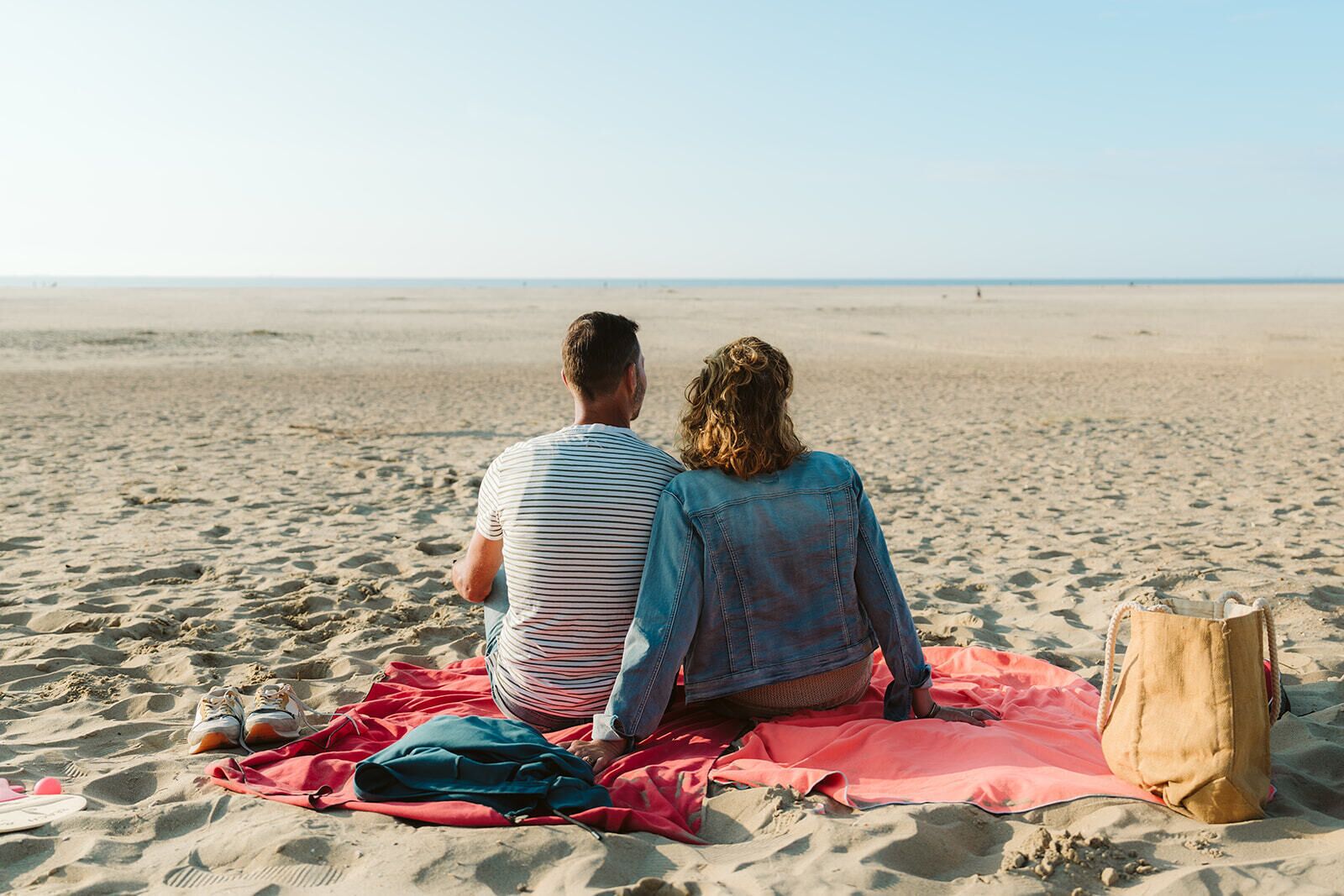 Zwei Personen sitzen auf einer Decke an einem sandigen Strand mit freiem Blick auf den Ozean.