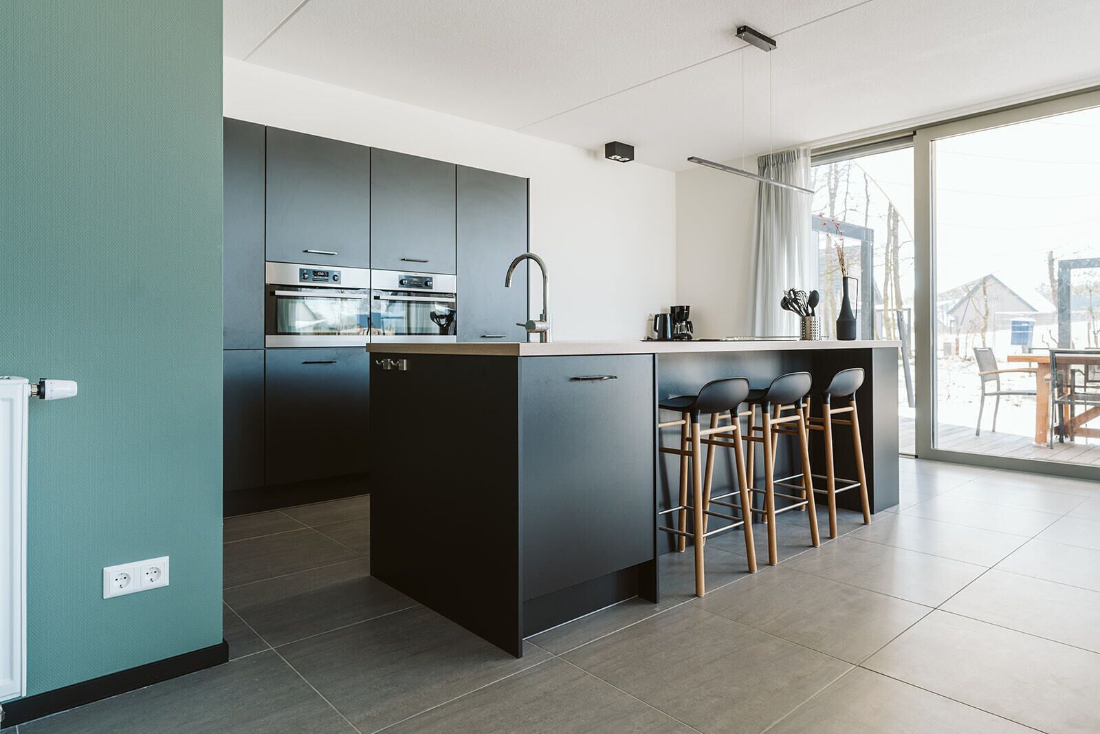 A modern kitchen with dark cabinetry, a central island with stools, and a large window providing a view of the outdoors.
