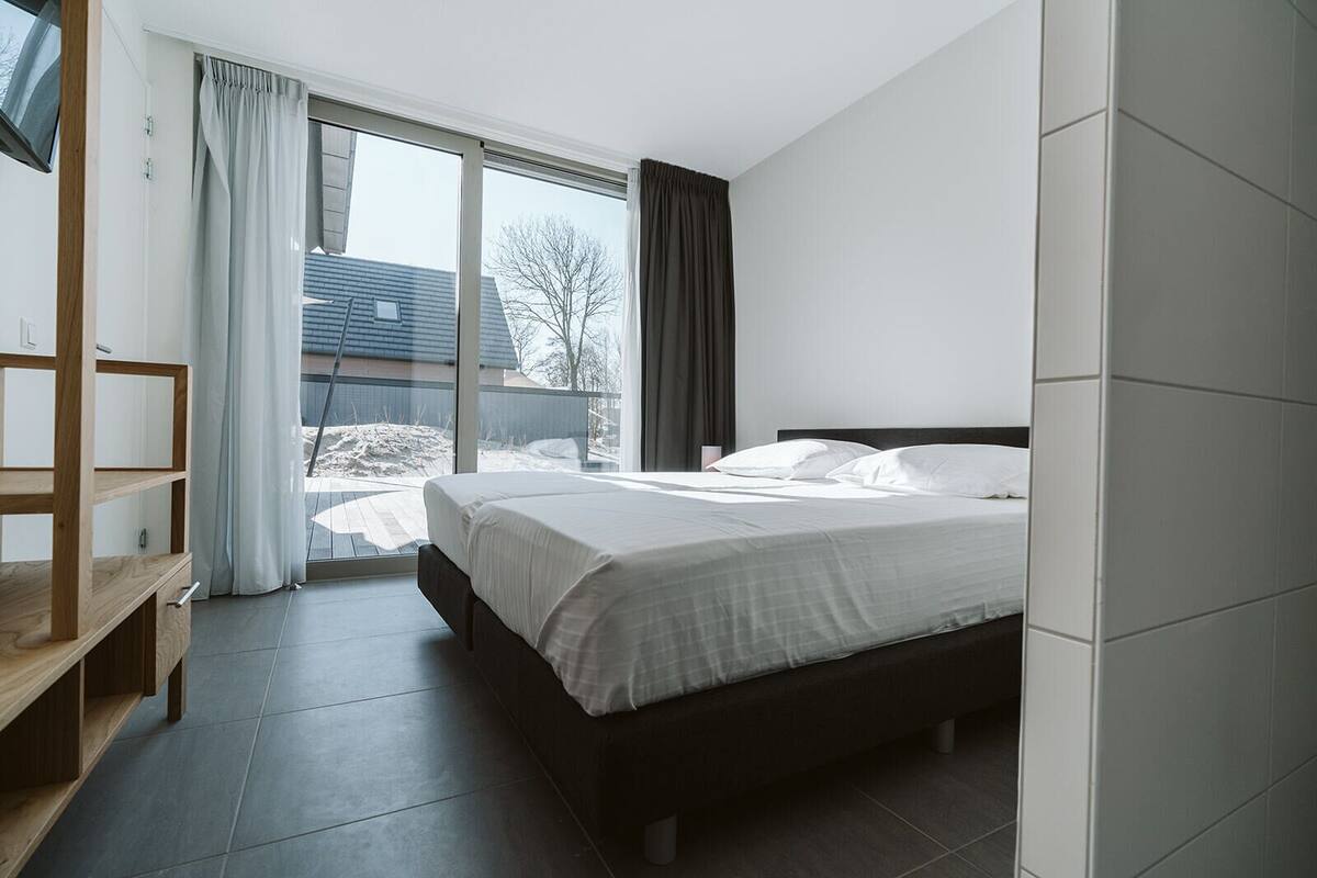 A modern bedroom with a large bed, a wooden shelf, and a view of the outside.