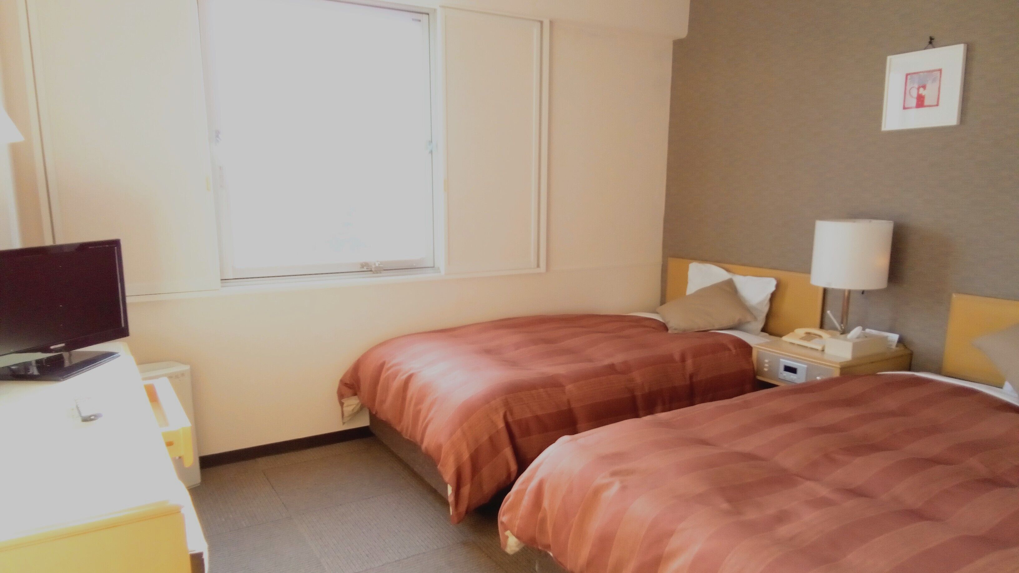 A hotel room with two beds, a television, a bedside table with a lamp, and a window.