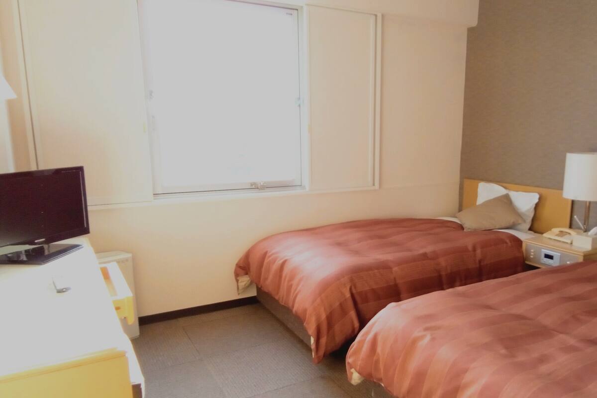 Ein Hotelzimmer mit zwei Betten, einem Fernseher, einem Nachttisch mit Lampe und einem Fenster.
