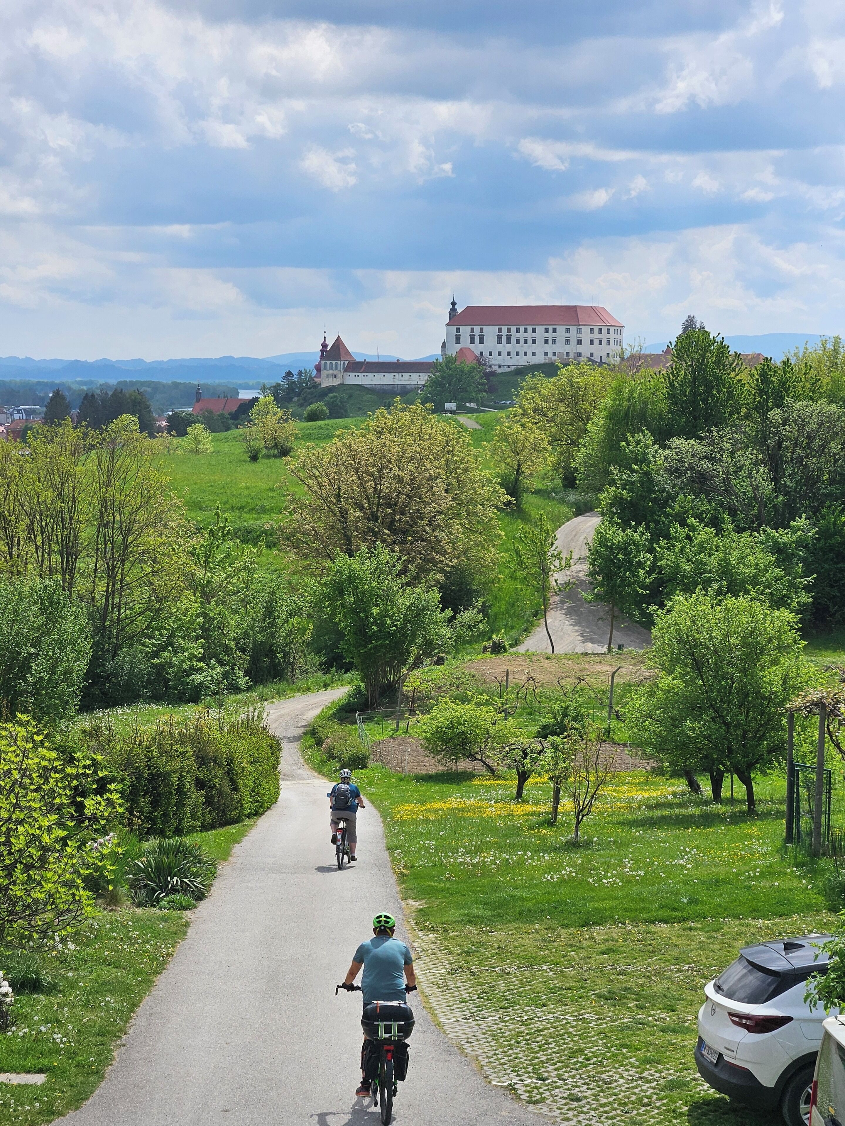 Une allée pavée où circulent des cyclistes, bordée de verdure et avec un grand bâtiment en arrière-plan.