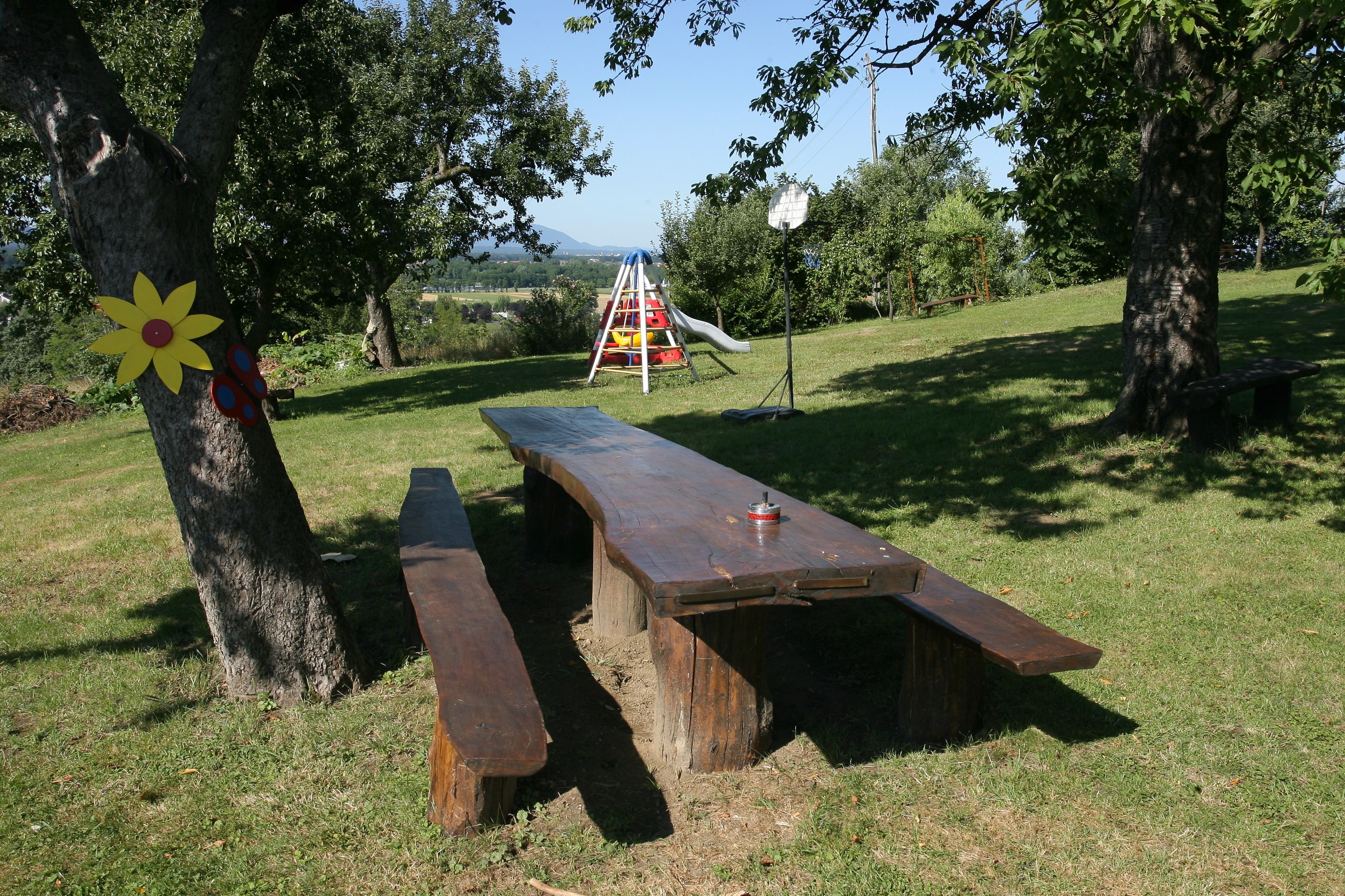 An outdoor picnic area with a wooden bench and picnic table, surrounded by trees and a playground in the background.