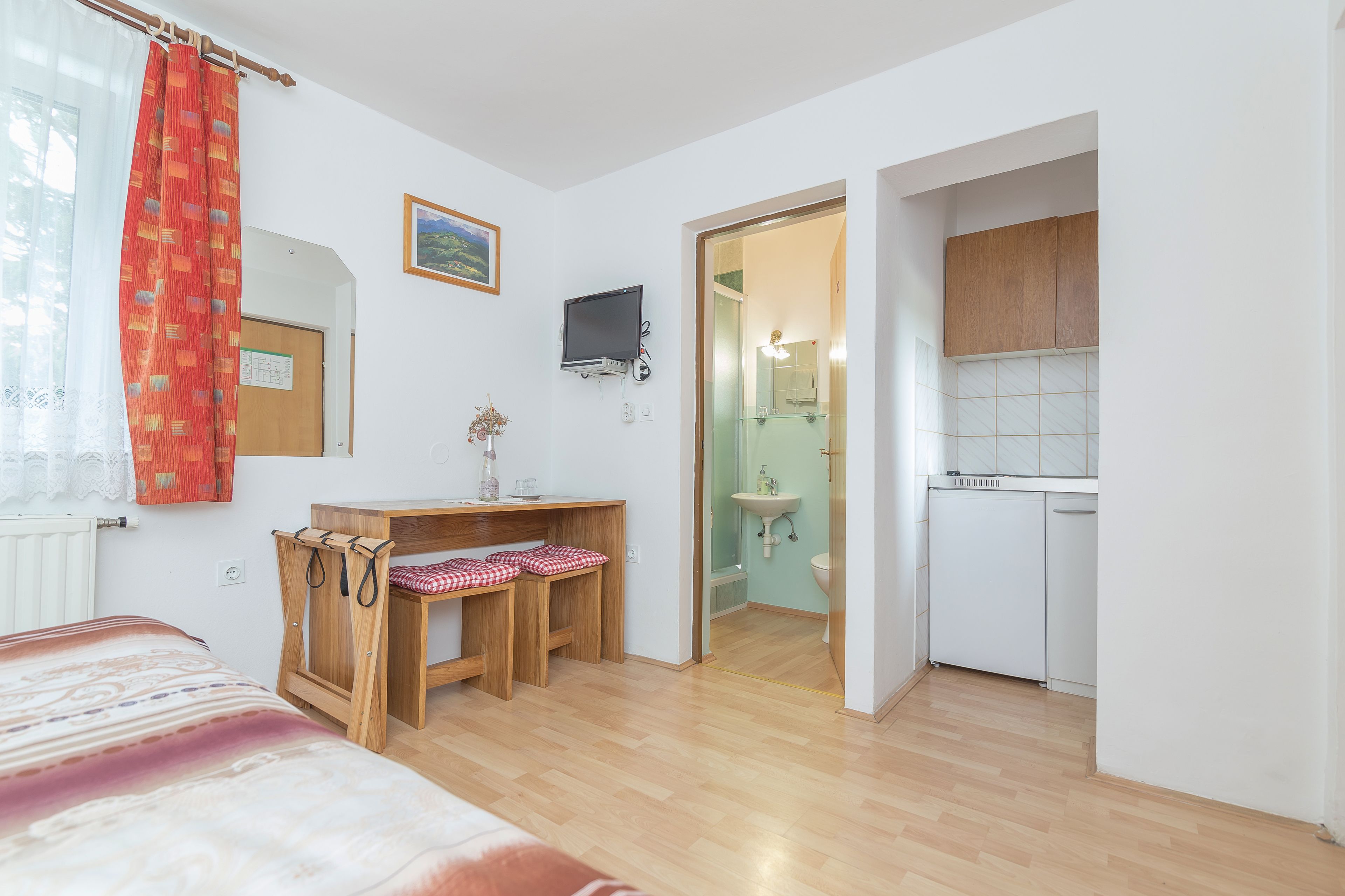 A bedroom with a bed, a desk with a chair, a TV, and a bathroom with a sink and toilet.
