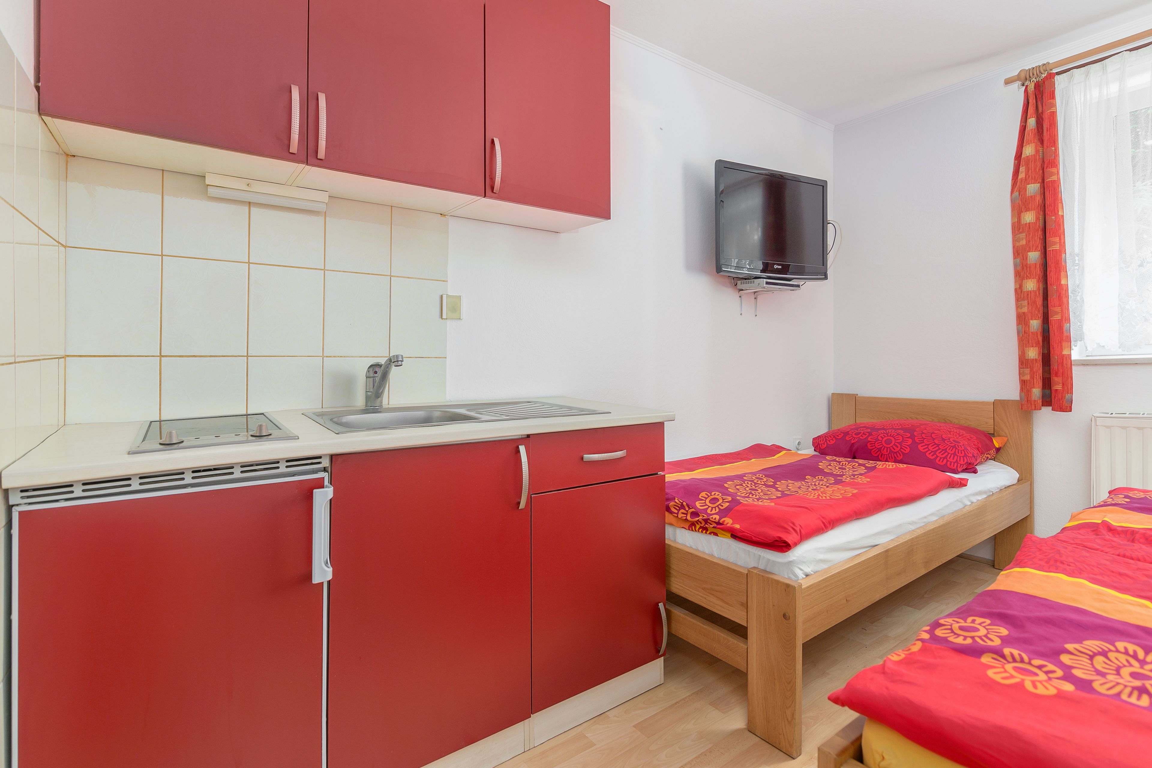 A compact room with a kitchenette, a bed with a red and orange bedspread, and a television mounted on the wall.
