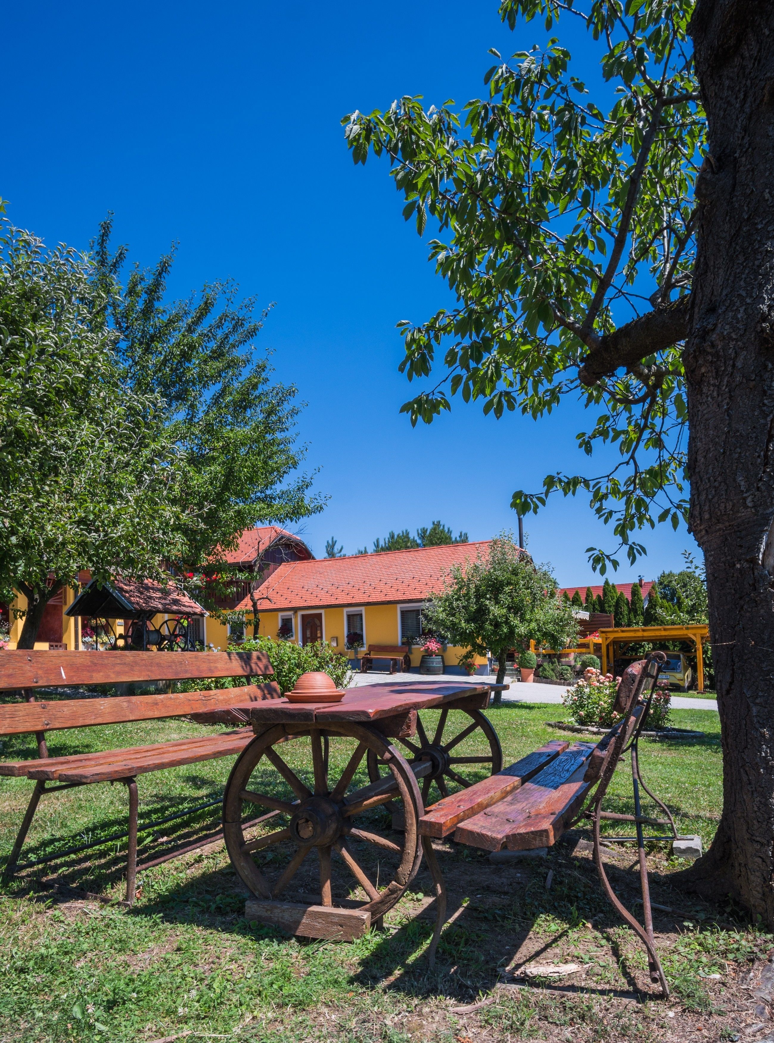 Un espacio al aire libre rústico con bancos de madera y una gran rueda de madera, rodeado de abundante vegetación y un edificio amarillo con tejado rojo.