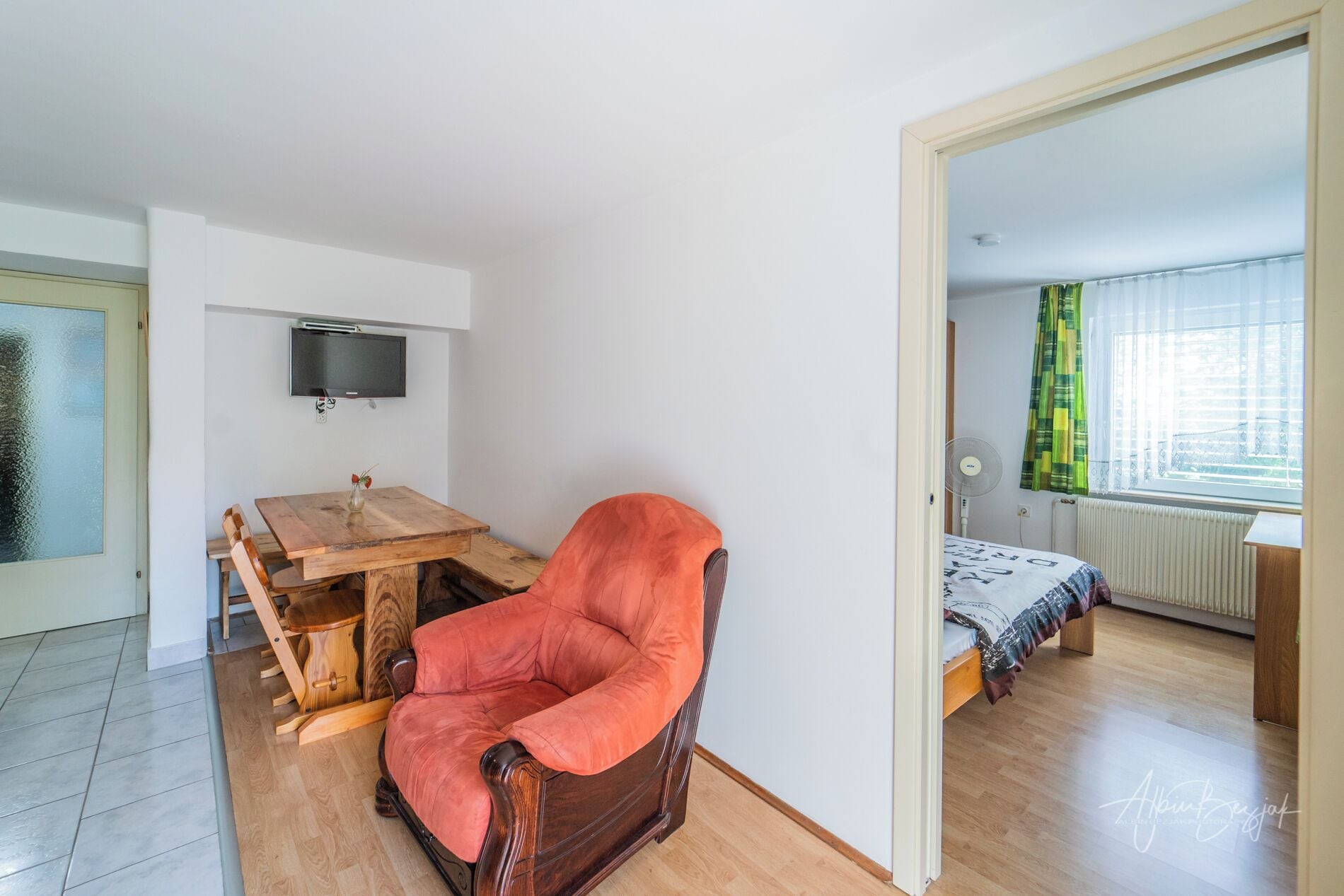Quarto com escrivaninha de madeira, uma cadeira vermelha, cama com colcha estampada, televisão fixada na parede e janela com cortinas.