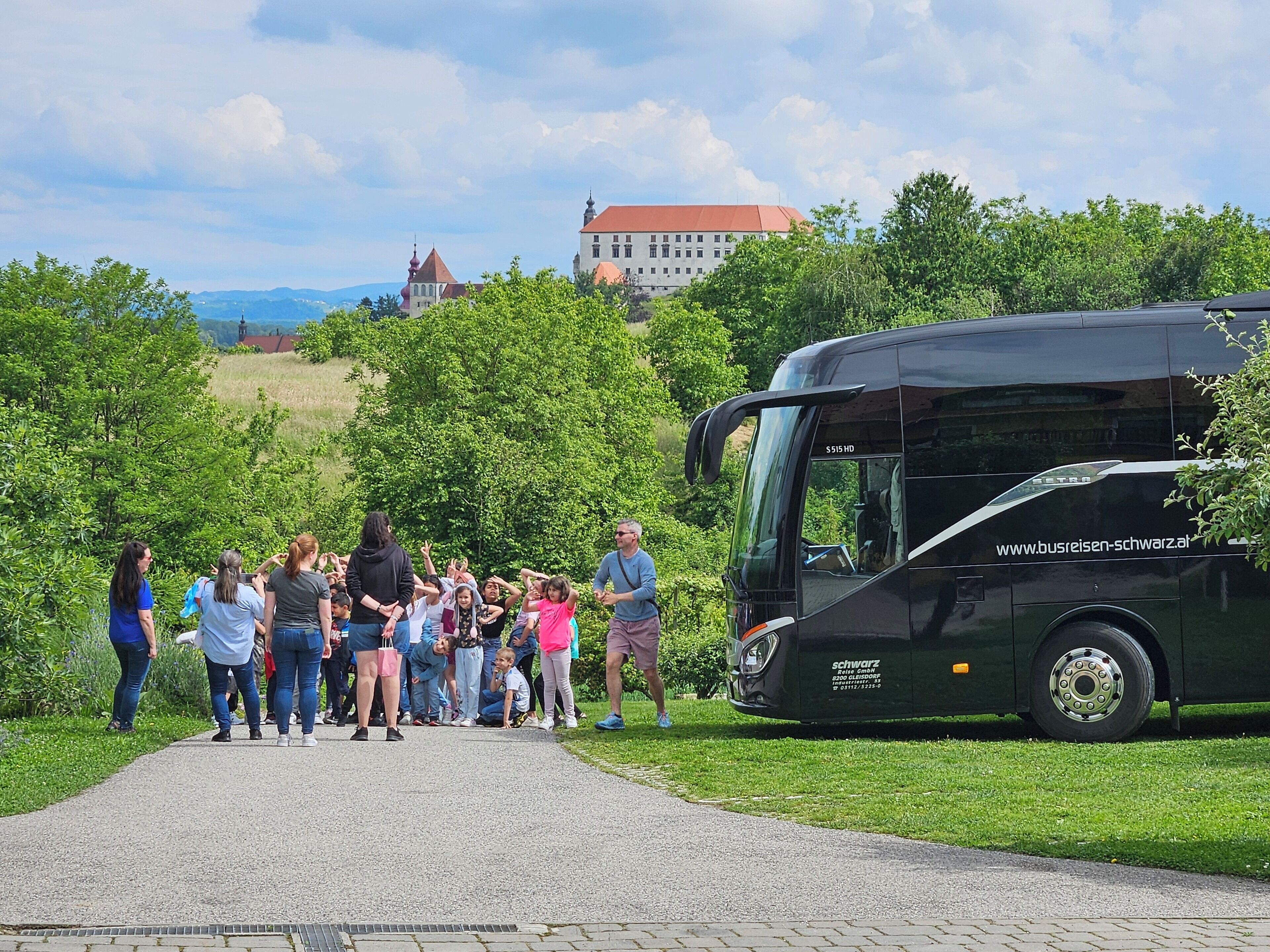 A group of people gathered near a bus, with a building and trees in the background.