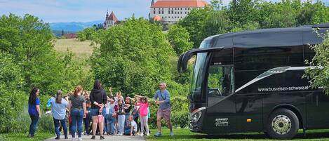 A group of people gathered near a bus, with a building and trees in the background.