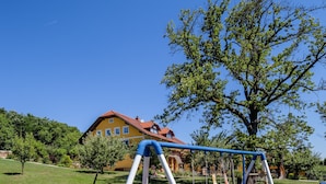 A playground with a swing set and slide, surrounded by grass and trees, with a house in the background. - Panorama B&B (Ptuj)