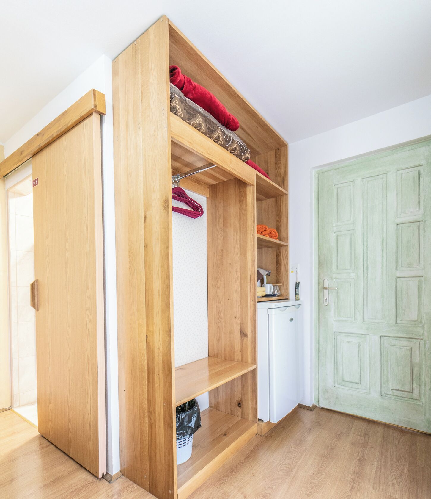Un armario de madera con varios estantes y espacio para colgar ropa, un refrigerador blanco y una puerta verde en una habitación con suelo de madera.