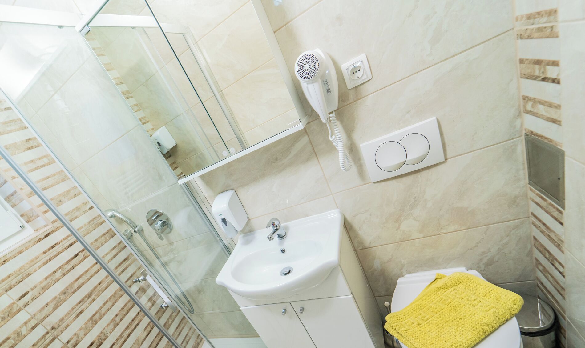 Un baño con cabina de ducha de vidrio, un lavabo blanco, un secador de pelo y una toalla amarilla.