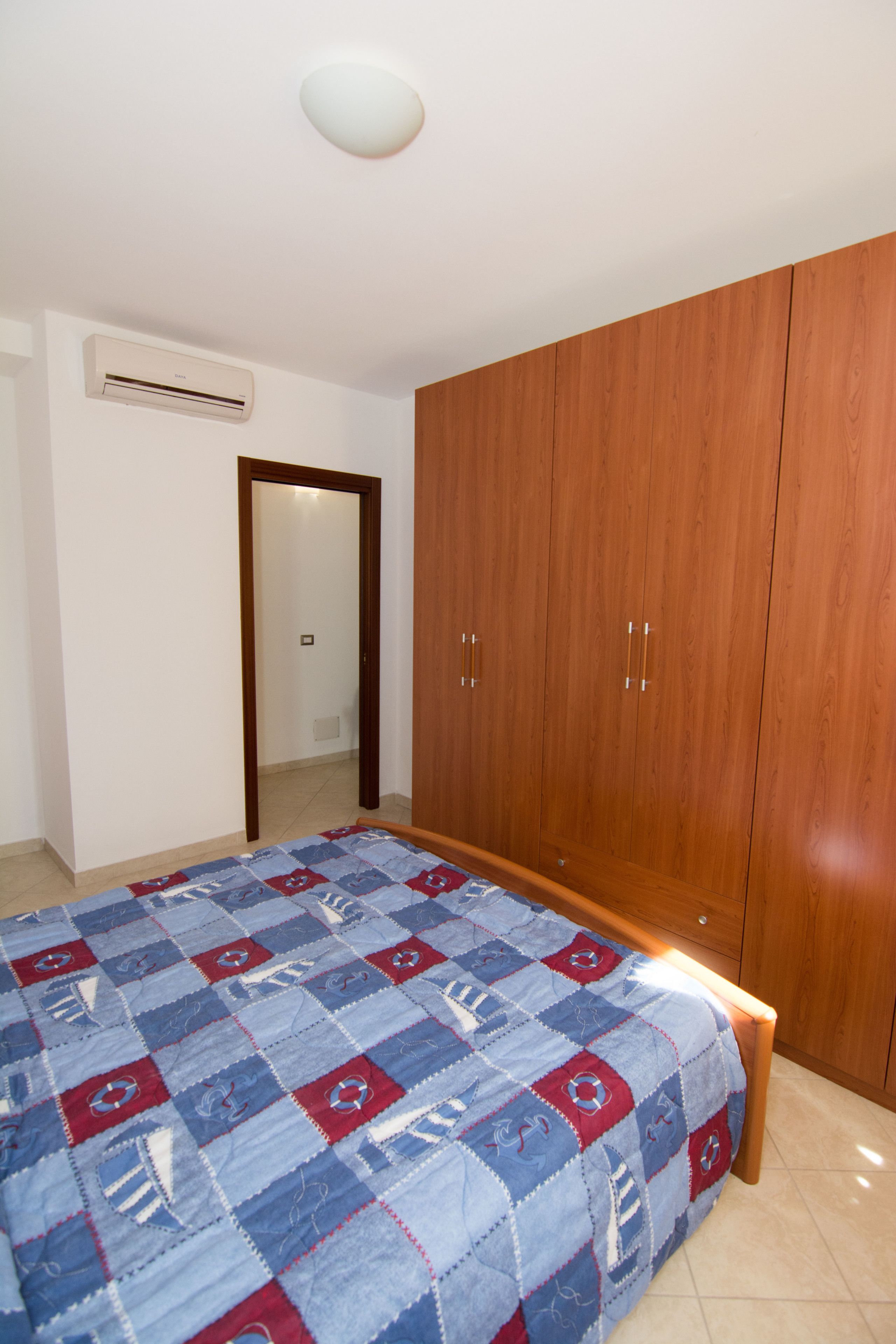 Une chambre avec une armoire en bois, un lit recouvert d’une couette bleue et rouge, et un climatiseur fixé au mur.