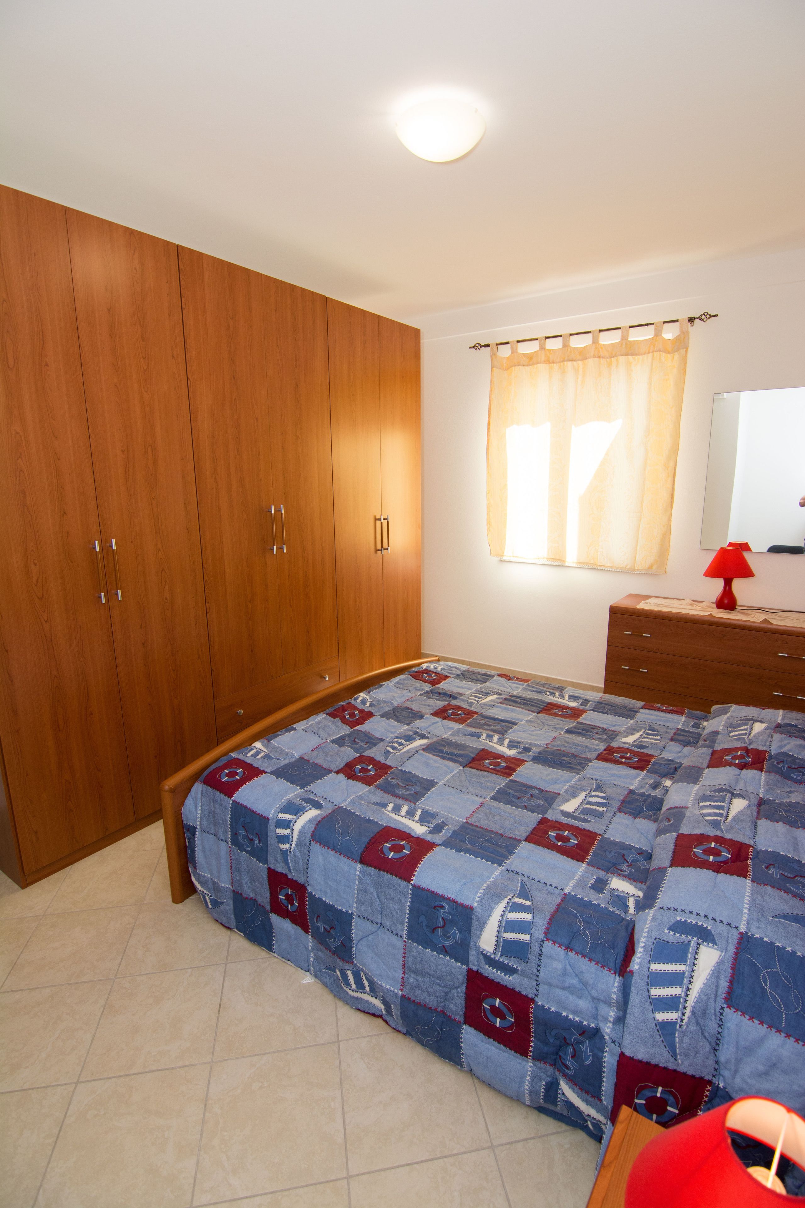 A bedroom with a wooden wardrobe, a bed with a quilt, a dresser, a mirror, and a window with curtains.