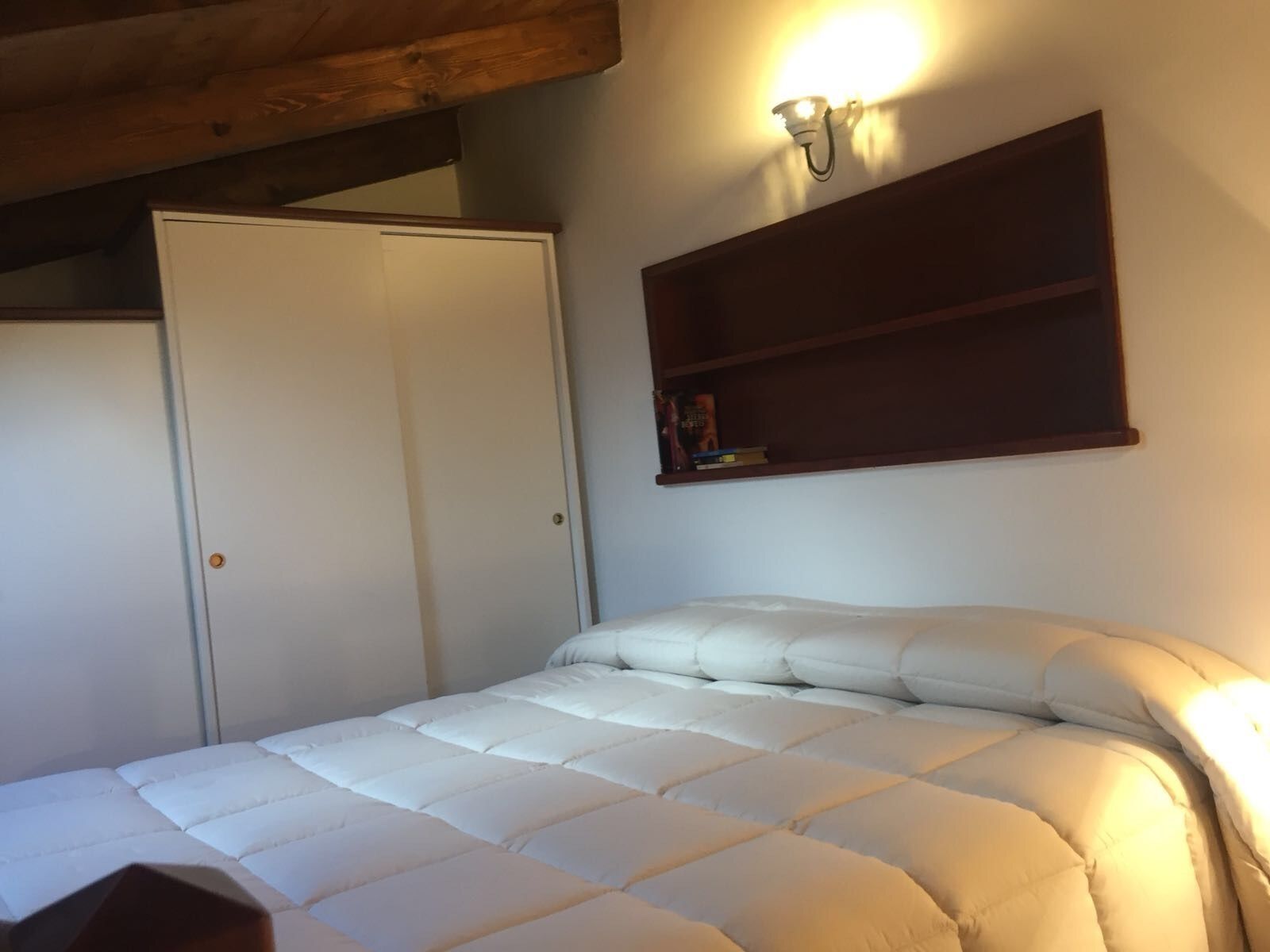 Een slaapkamer met een wit bed, een houten plafond, een witte deur en een wandplank met boeken.
