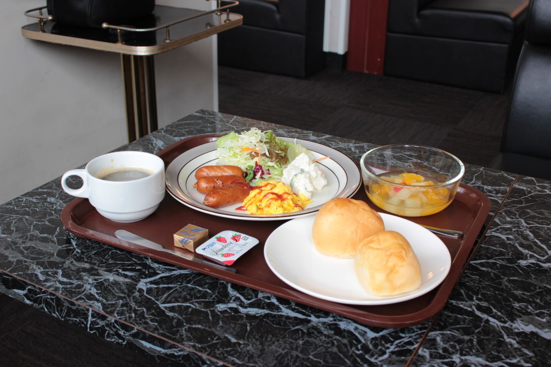 A tray with breakfast items including sausages, scrambled eggs, a salad, a bowl of fruit, a cup of coffee, and a small packet of butter.
