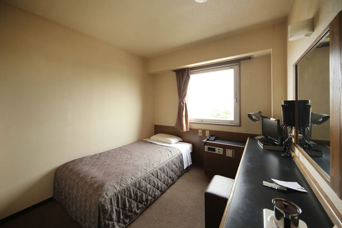 A small hotel room with a single bed, a desk with a telephone and a mirror, and a window with curtains.