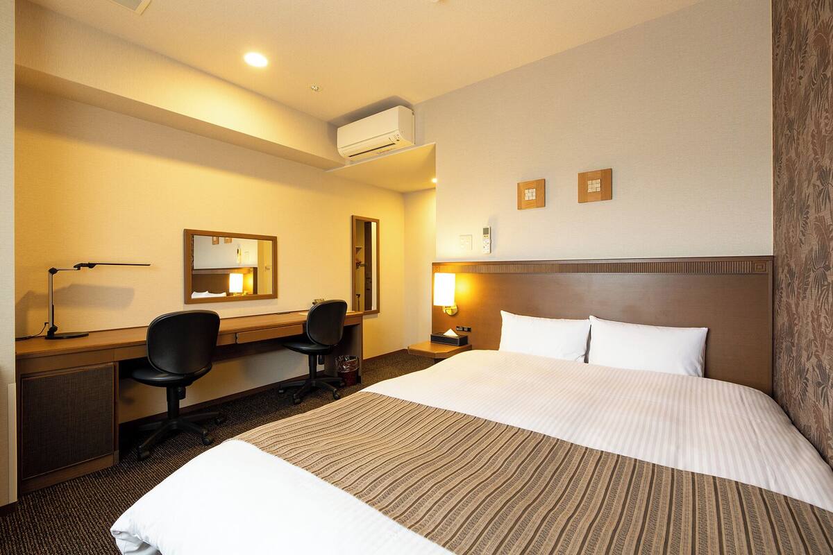 A hotel room with a bed, desk, chair, and a wall-mounted air conditioner.