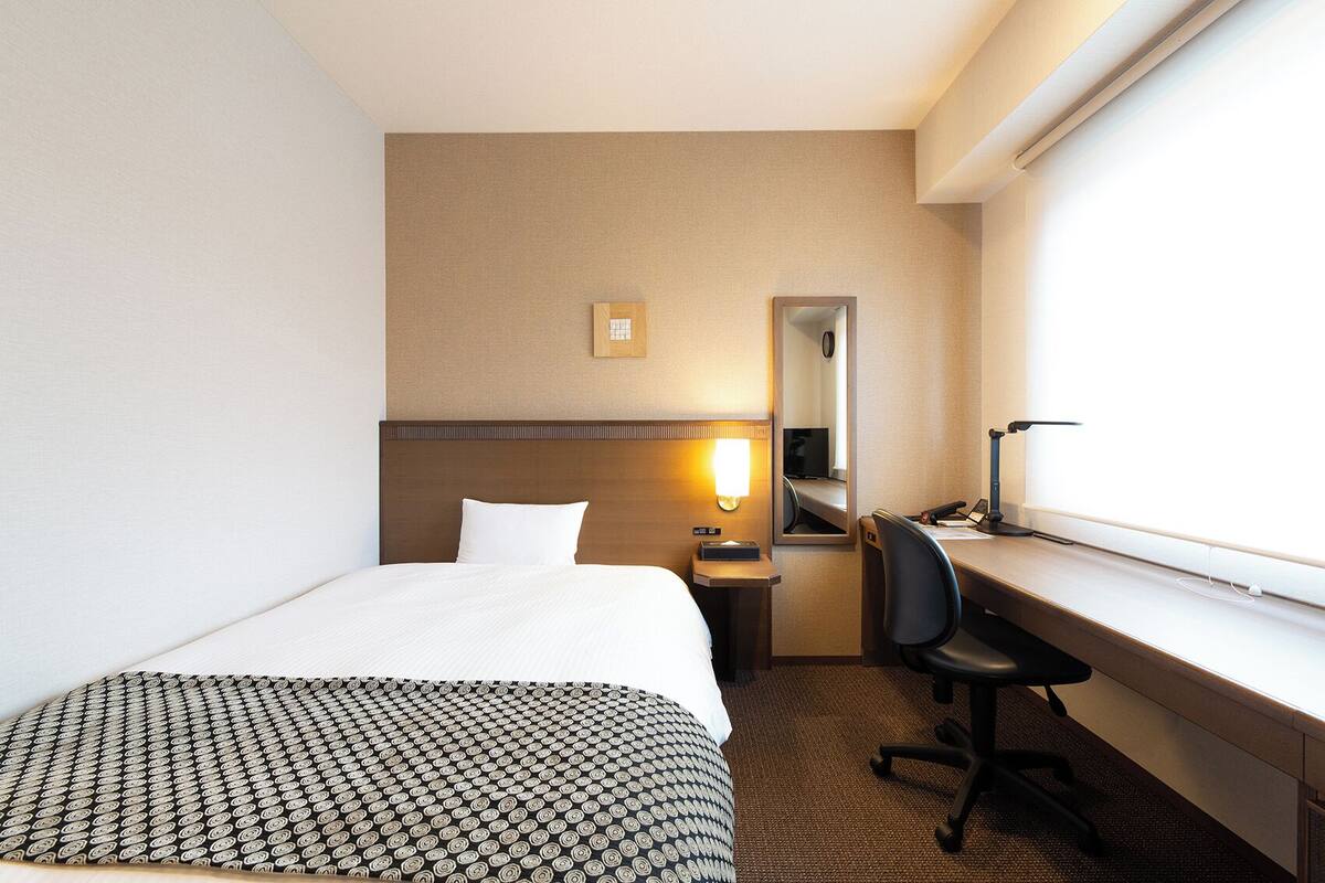 A hotel room with a bed, a desk, a chair, a desk lamp, a mirror, and a wall-mounted light.