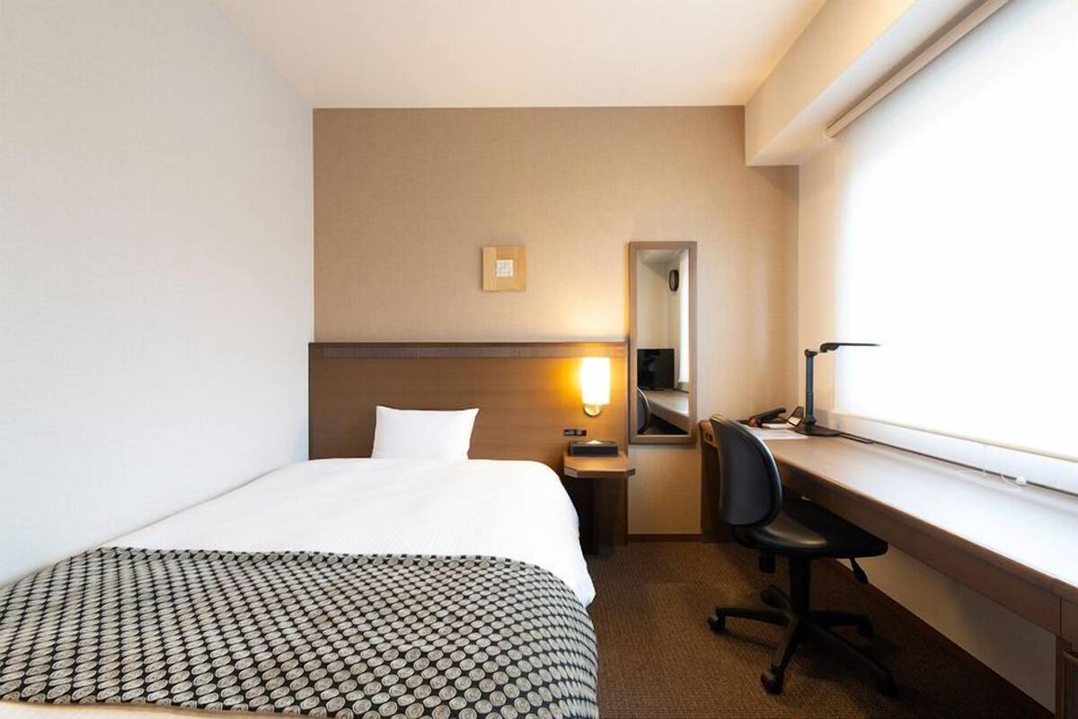 A hotel room with a bed, desk, chair, and a window.