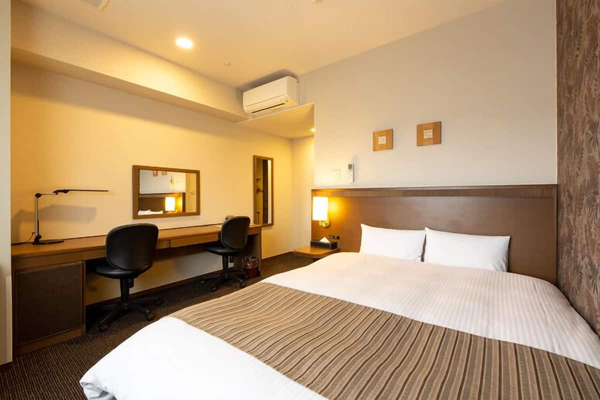 A hotel room with a large bed, a desk with a chair, a desk lamp, a wall-mounted air conditioner, and a nightstand with a lamp.