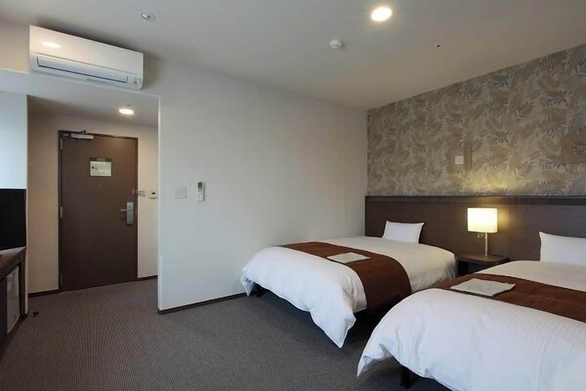 A hotel room with two beds, a wall-mounted air conditioner, a door with a sign, and a nightstand with a lamp.